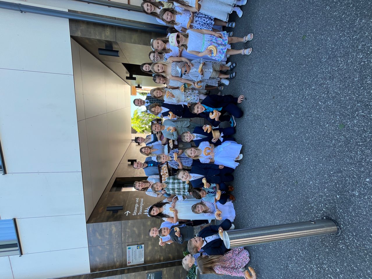 Eine große Gruppe von Kindern steht draußen, hält Donuts in der Hand und posiert für ein Foto. Sie stehen auf einem Bürgersteig in der Nähe eines Gebäudes.