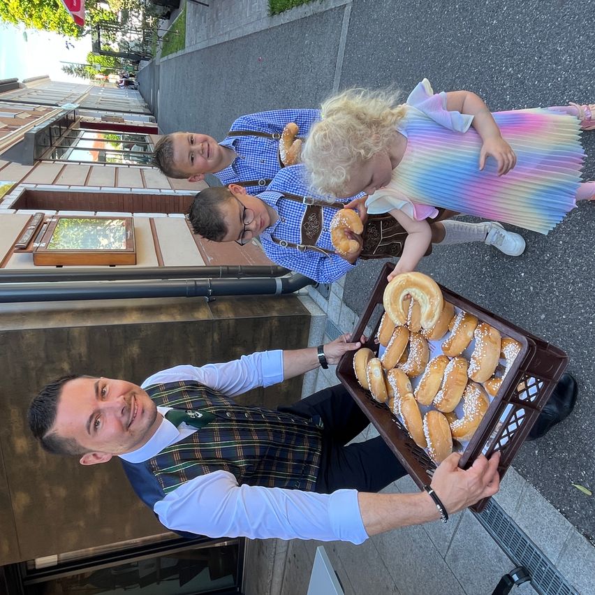 Ein Mann hält einen Korb mit Donuts, während zwei Kinder hinter ihm stehen. Ein Kind hält einen Donut, während das andere in die Kamera schaut. Die Szene spielt sich vor einem Gebäude ab.