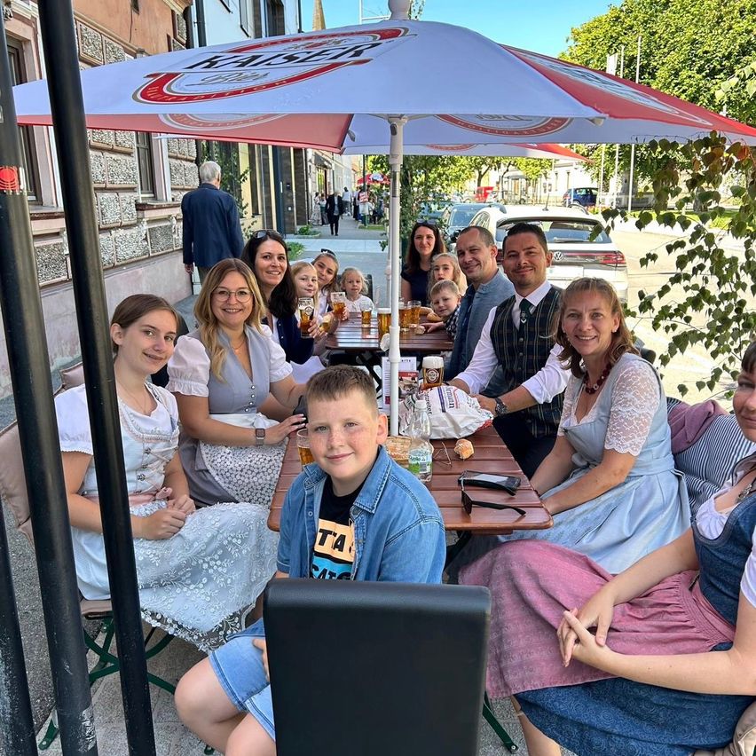 Eine Gruppe von Menschen sitzt um einen Tisch in einem Café im Freien und lächelt für die Kamera. Sie sind in traditioneller bayerischer Kleidung gekleidet, und einige haben Gläser mit Bier. Ein Schirm mit dem Wort Kaiser steht über ihnen.