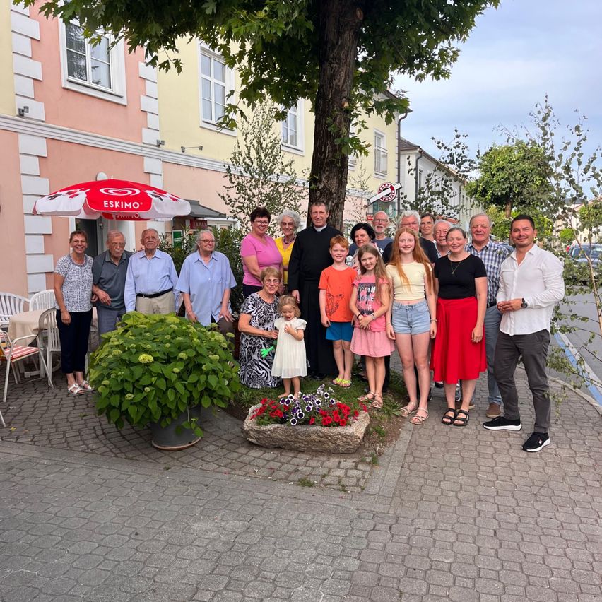 Eine Gruppe von Menschen posiert für ein Foto vor einem Baum und Blumen. Ein Mann steht in einem schwarzen Gewand. Sie befinden sich in einem Außenbereich mit Gebäuden und Autos im Hintergrund.