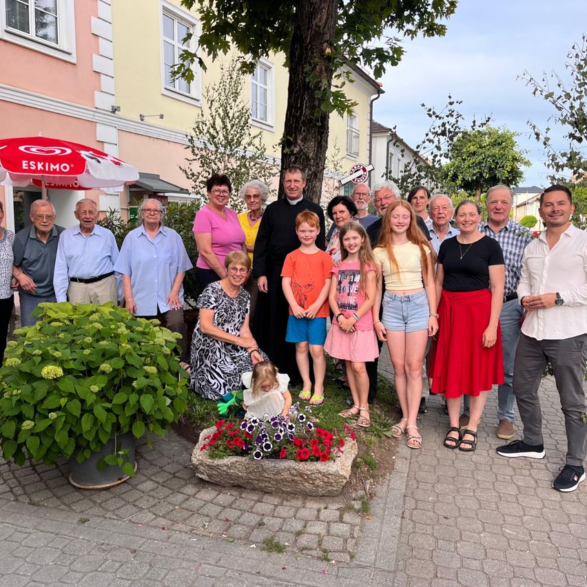 Eine große Gruppe von Erwachsenen und Kindern posiert für ein Foto vor einem Baum, mit Gebäuden und einem roten Schirm im Hintergrund.
