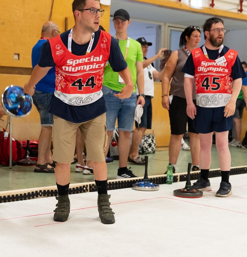 Bei den Special Olympics treten Athleten in verschiedenen Sportarten an, darunter auch im Curling. Hier bereiten sich zwei Athleten darauf vor, ihre Curlingsteine auf einer Bahn zu werfen.