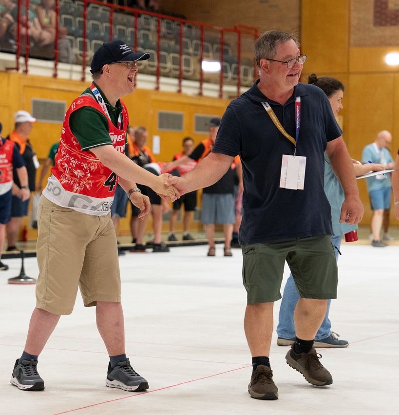 Zwei Männer schütteln sich die Hände bei einem Sportereignis. Einer trägt ein rot-weißes Shirt mit der Nummer 4. Der andere trägt ein blaues Shirt und grüne Shorts. Sie stehen auf einem markierten Platz mit anderen im Hintergrund.