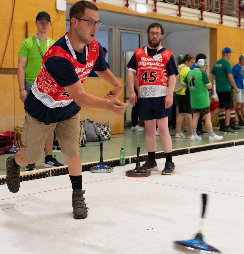 Ein Mann in einer Special Olympics-Uniform bereitet sich darauf vor, einen Curlingstein auf einer Eisbahn zu werfen, während ein anderer Mann hinter ihm steht. Zuschauer beobachten von den Seitenlinien.