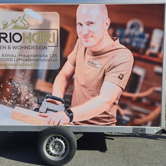 Ein Mann in einem braunen T-Shirt sägt ein Stück Holz mit einer Kreissäge. Dahinter steht ein Holzregal mit Werkzeugen und Geräten.