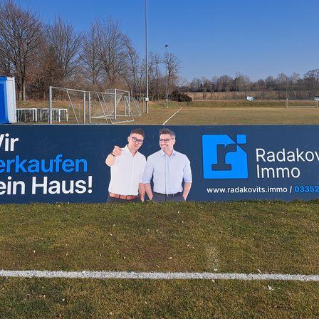 Ein großes blau-weißes Werbebanner steht auf dem Rasenfeld mit zwei Männern davor. Es steht geschrieben: 'Wir verkaufen ein Haus!' und zeigt das Firmenlogo.