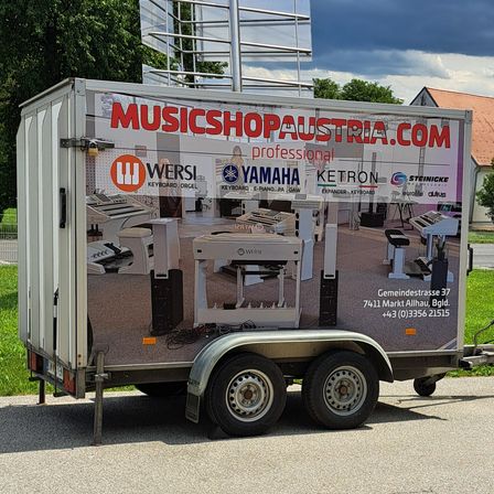 Ein weißer Anhänger für MUSICSHOPAUSTRIA.COM auf einer Straße geparkt. Der Anhänger zeigt ein Bild eines Klaviers und anderer Musikinstrumente auf seiner Seite.