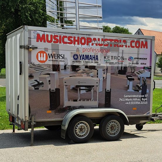 Ein weißer Anhänger für MUSICSHOPAUSTRIA.COM auf einer Straße geparkt. Der Anhänger zeigt ein Bild eines Klaviers und anderer Musikinstrumente auf seiner Seite.