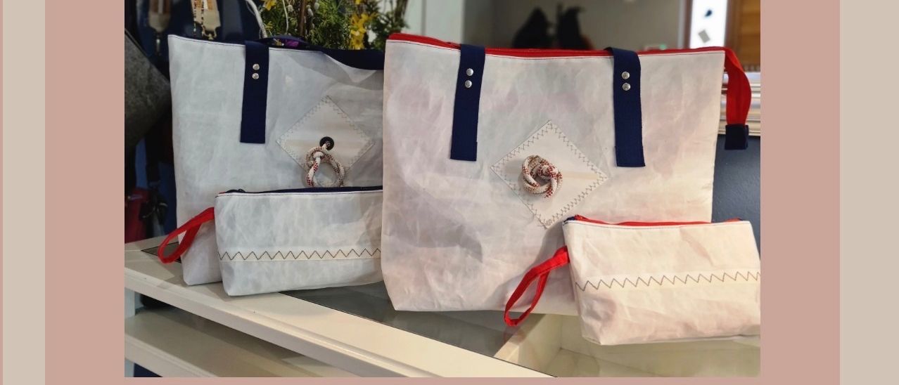 Drei weiße Taschen mit roten und blauen Henkeln und ein kleiner Beutel mit einem Seil liegen auf einem Tisch. Die Taschen haben weiße und rote Aufnäher. Hinter den Taschen steht eine Pflanze mit gelben Blüten.