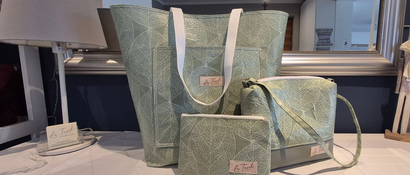 Drei Taschen mit Blattmustern sind auf einer weißen Oberfläche ausgestellt. Eine große Tasche, eine mittlere Tasche und eine kleine Handtasche sind zu sehen. Alle haben weiße Griffe.