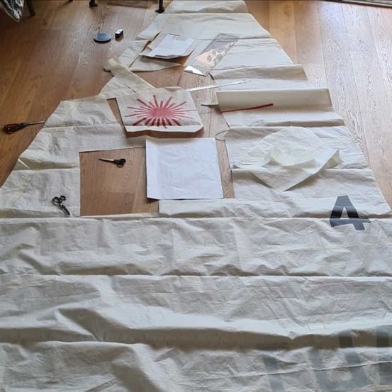 Ein Holzboden mit einem großen weißen Tuch ausgebreitet. Das Tuch hat die Nummer '4' und ein rotes Sonnendesign darauf. Werkzeuge und andere Gegenstände sind auf dem Boden verstreut.