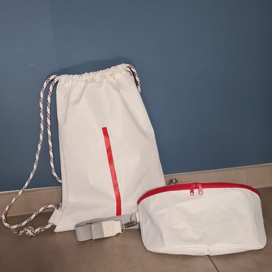 Eine weiße Tasche mit Kordelzug und rotem Streifen sowie eine kleinere Tasche mit Reißverschluss stehen auf einem gefliesten Boden an einer blauen Wand.