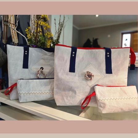 Drei weiße Taschen mit roten und blauen Griffen auf einem Glasregal, jeweils mit dekorativer Stickerei. Im Hintergrund eine Blumenarrangement.