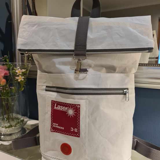 Ein weißer Rucksack mit grauem Riemen und rotem Etikett, das 'Laser Neue Nummern 3-8' sagt. Der Rucksack ist auf einer Glasfläche mit einem Blumenvase im Hintergrund ausgestellt.