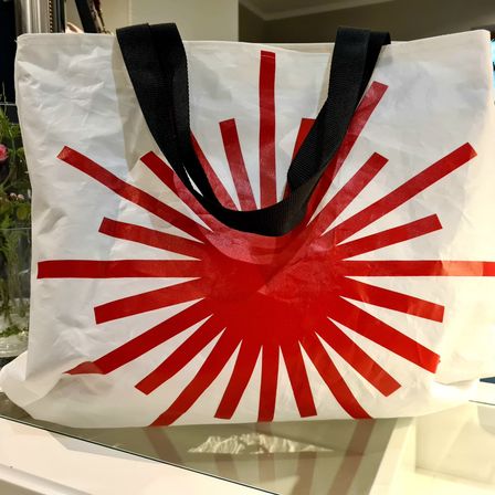 Ein weißer Tragetasche mit rotem Sonnenaufgangsdesign und schwarzen Henkeln wird auf einer Glasoberfläche präsentiert.