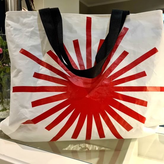 Ein weißer Tragetasche mit rotem Sonnenaufgangsdesign und schwarzen Henkeln wird auf einer Glasoberfläche präsentiert.