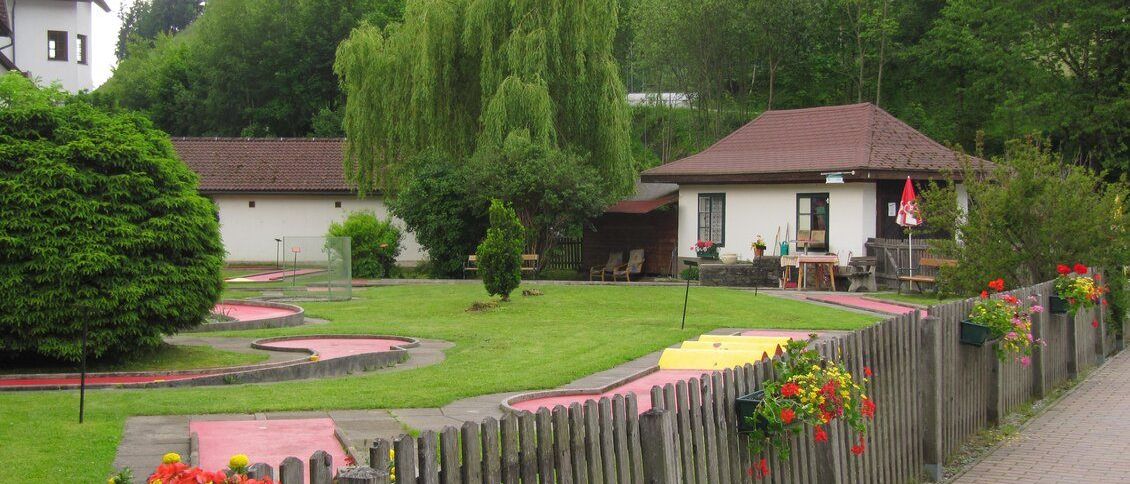 Ein Minigolfplatz mit einem Holzzaun. Im Hintergrund befinden sich Bäume, ein Haus und eine Bank. Das Haus hat eine rote Flagge und einen geschlossenen Regenschirm.