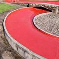 Zwei Männer spielen Minigolf auf einem Platz mit einem rot gestrichenen Loch, in der Nähe eines Hauses mit braunem Dach und einer Bank.