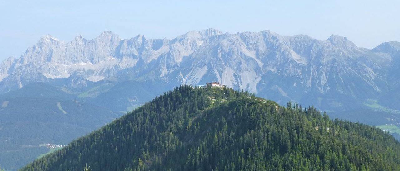 Ein Panoramablick auf eine Bergregion mit einem kleinen Haus auf einem Hügel unter einem klaren Himmel.