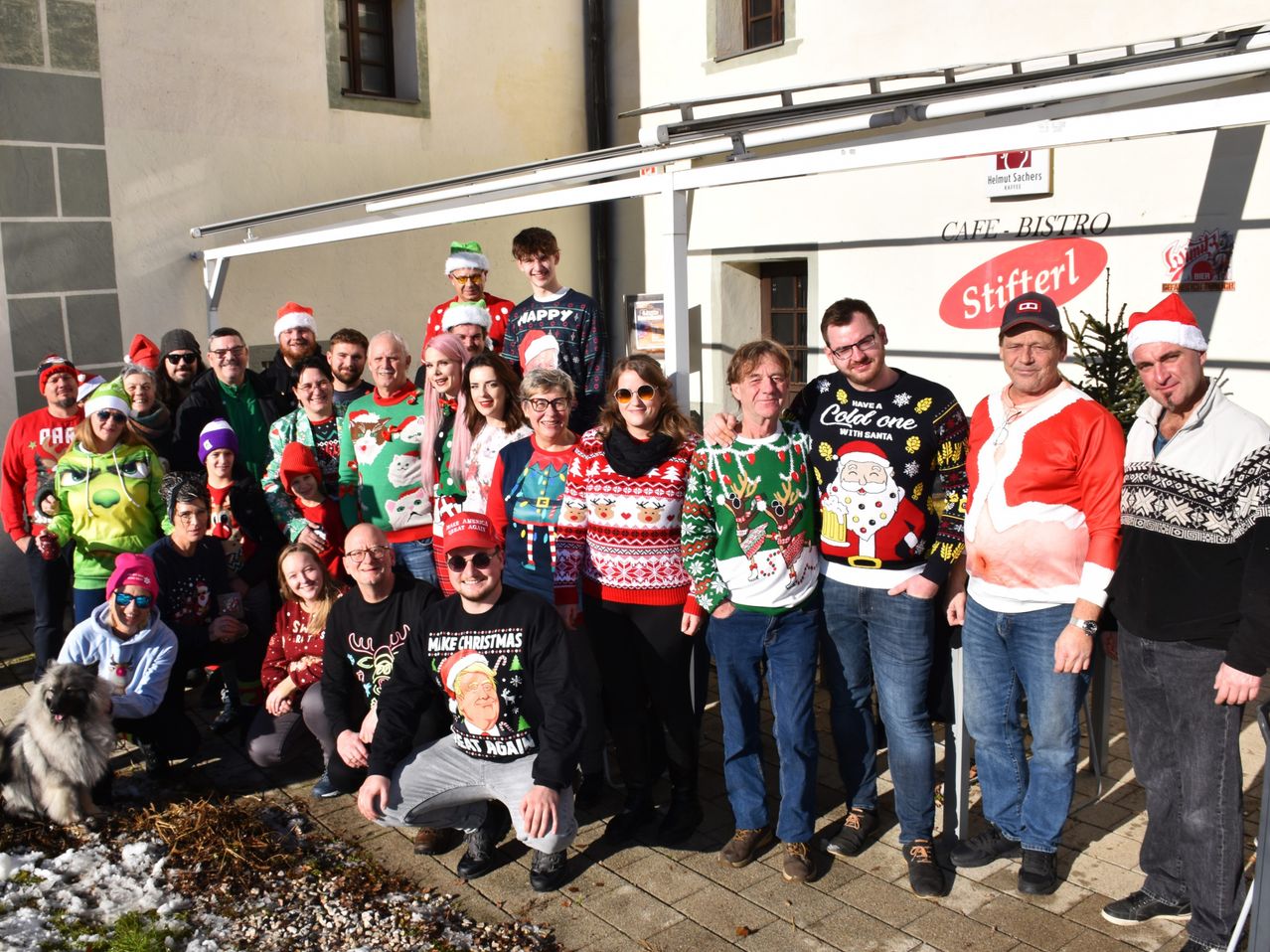 A group of people wearing Christmas sweaters poses for a photo in front of a building. Some are wearing hats and glasses. The man in the middle wears a Santa Claus sweater.