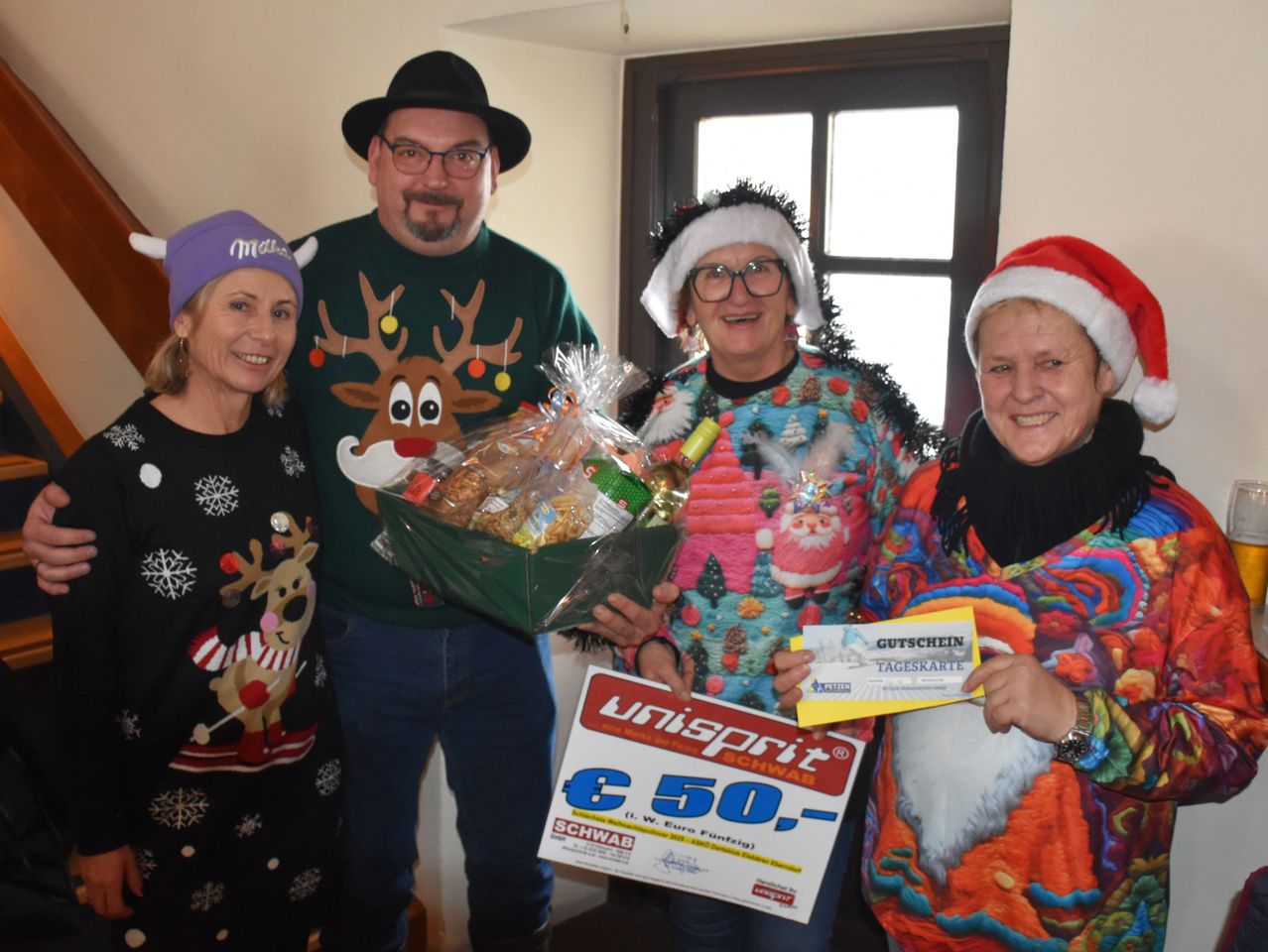 Vier Erwachsene in Weihnachtspullovern und Mützen posieren für ein Foto. Ein Mann hält einen Geschenkkorb und ein Schild mit 50 €. Ein anderer hält eine Geschenkkarte. Sie stehen drinnen in der Nähe eines Fensters.