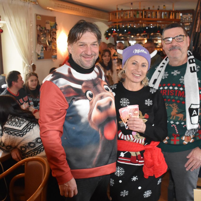 Drei Erwachsene lächeln in einem Raum mit Weihnachtsdekoration. Ein Mann trägt ein Rentier-Shirt. Eine Frau hält Geld und einen Schal. Ein Mann trägt eine Brille.