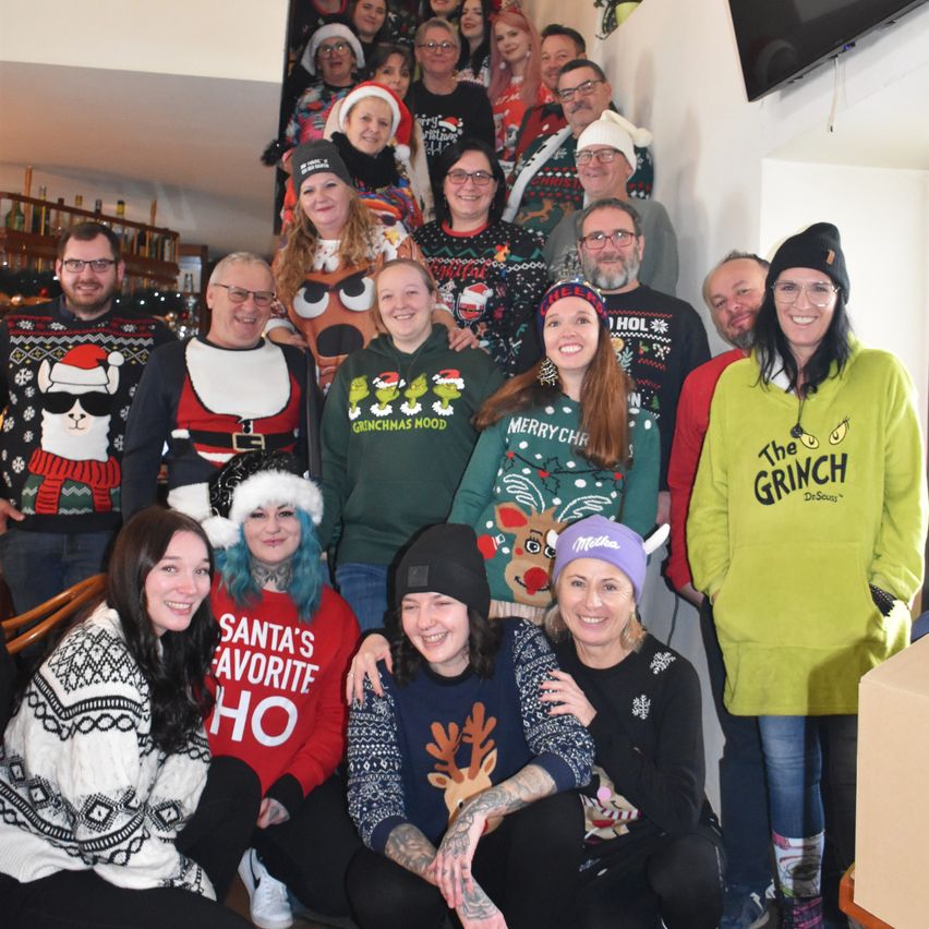 Eine Gruppe von Menschen in festlichen Pullovern und Mützen posiert für ein Foto. Einige tragen Weihnachtsmützen, Grinch-Hoodies und andere haben witzige Designs. Sie stehen in einem Raum mit einer Treppe im Hintergrund.