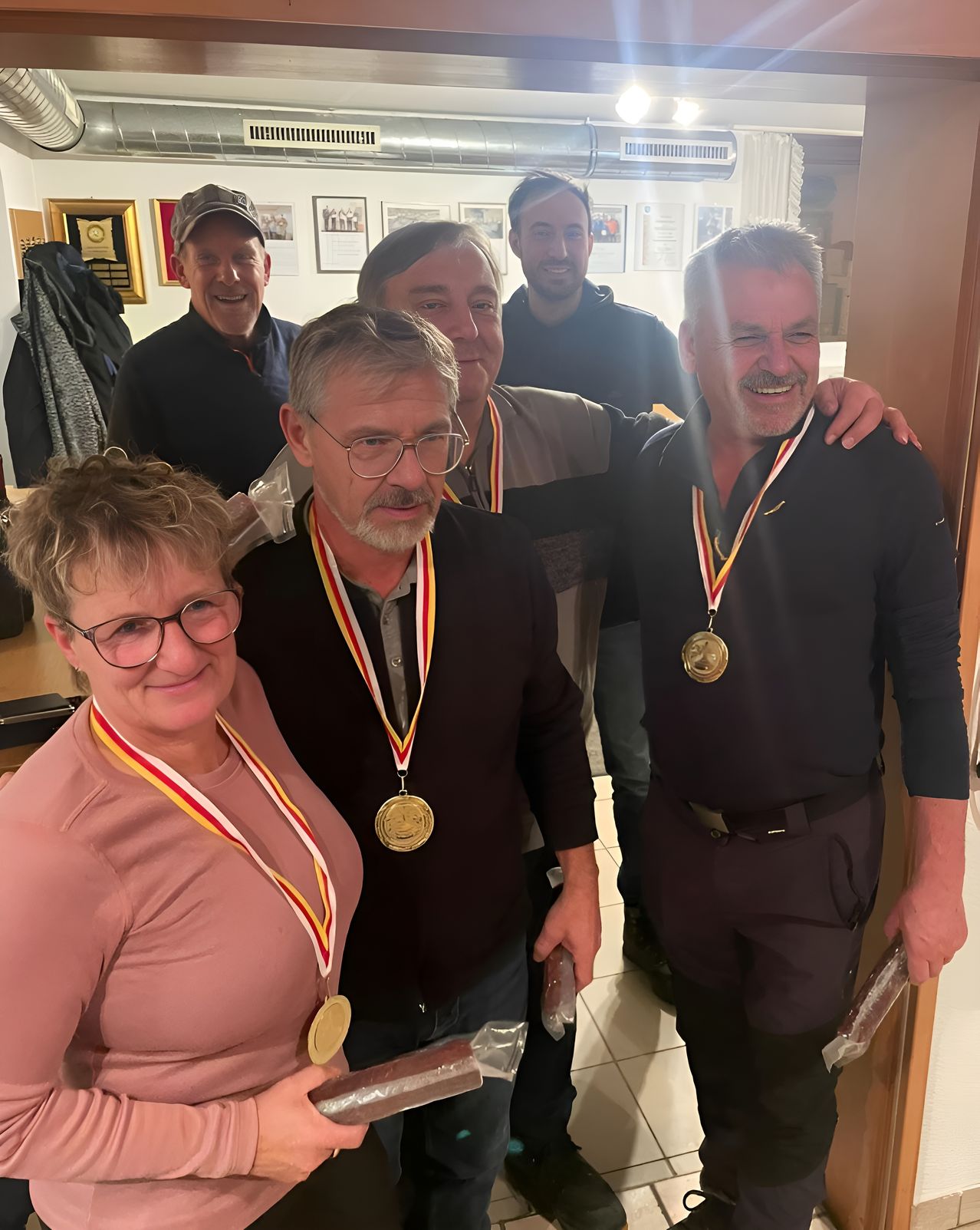 Eine Gruppe von Menschen lächelt und posiert für ein Foto in einem Raum. Sie tragen Medaillen und einige von ihnen halten Papiere. Der Mann auf der linken Seite trägt eine Brille und einen Hut. Hinter ihnen hängen Kleidungsstücke an der Wand.