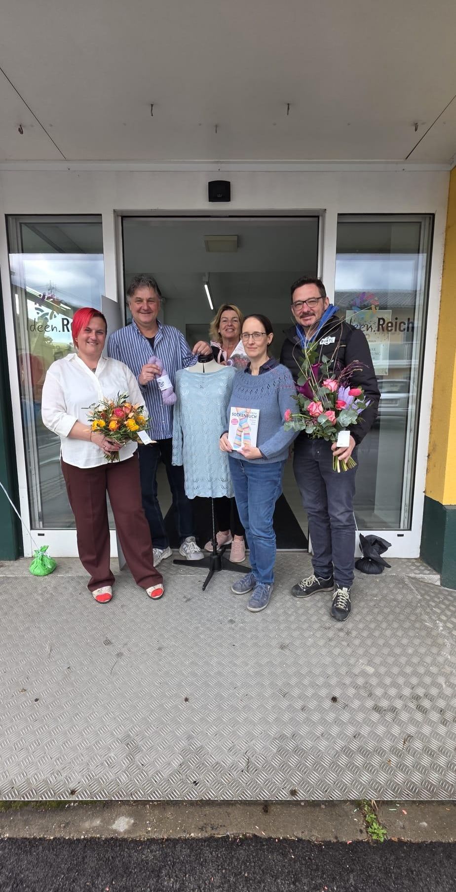 Fünf Personen stehen vor einem Geschäft mit Blumen und einem Buch. Einer hält ein gestricktes Stück, und ein anderer hält ein Mannequin mit einem blauen Kleid. Das Geschäftsschild lautet 'deen.Reich'.