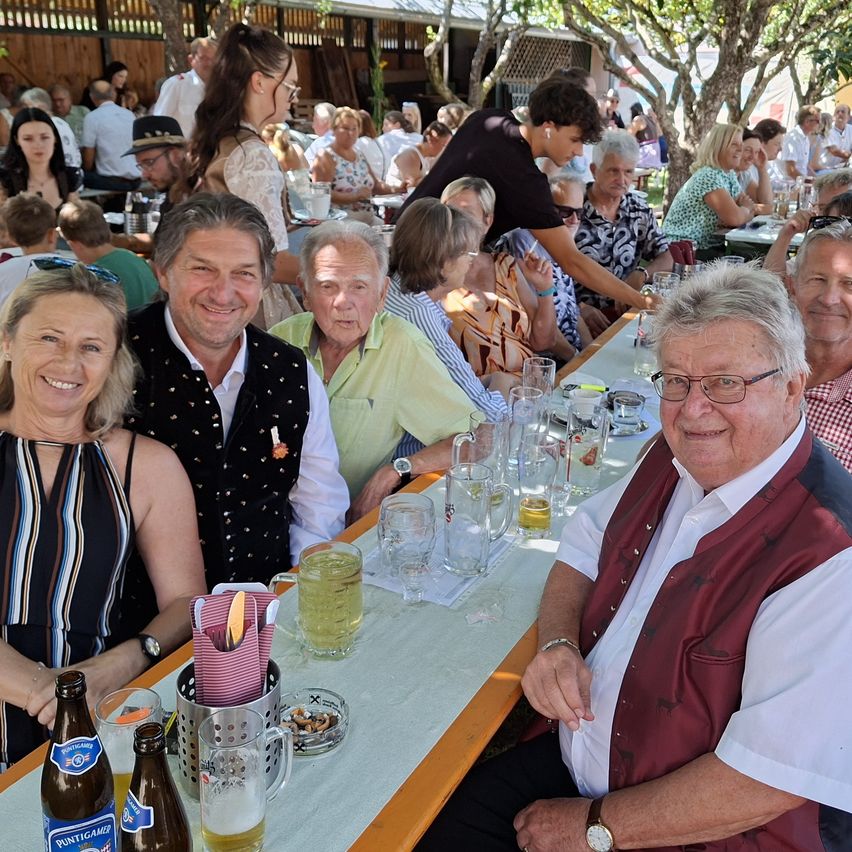 Eine Gruppe von Menschen sitzt an einem Tisch in einem Outdoor-Restaurant, lächelt und posiert für ein Foto. Der Tisch ist mit Gläsern, Flaschen und Servietten gefüllt. Im Hintergrund sind weitere Menschen zu sehen, die sitzen und stehen, einige mit Hüten.