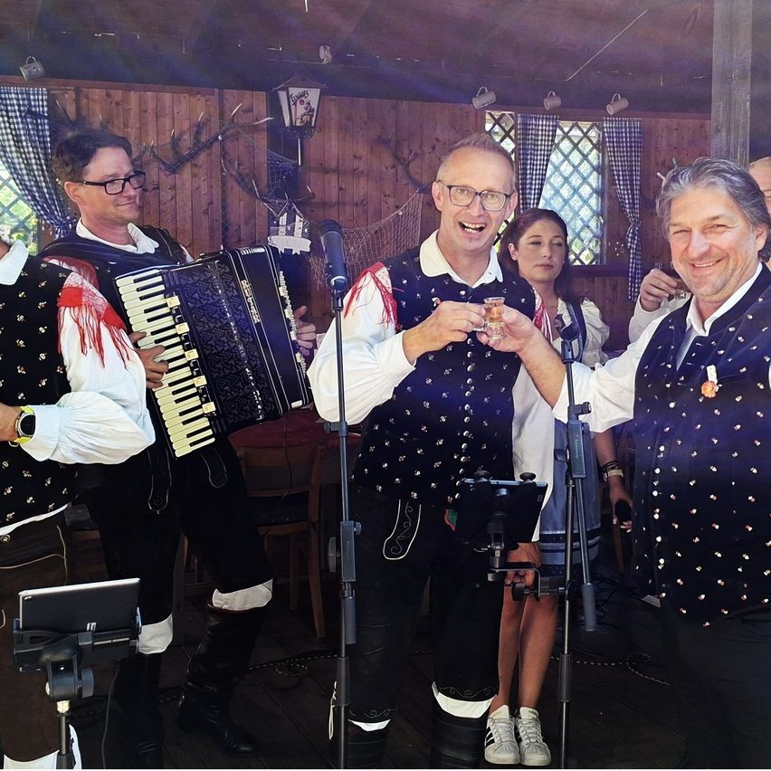Vier Männer in traditioneller Kleidung spielen Musik, einer spielt Akkordeon, zwei halten Getränke und der letzte lächelt. Eine Frau steht hinter ihnen und ein Mikrofon ist aufgestellt.