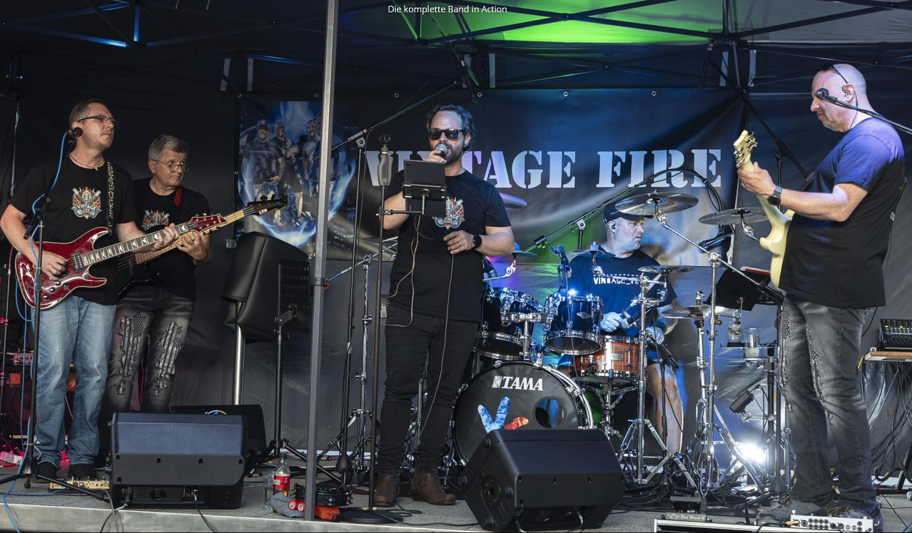 Eine Band spielt auf der Bühne, hinter ihnen ein Banner mit der Aufschrift 'Vintage Fire'. Der Leadsänger hält ein Mikrofon, während der Schlagzeuger und Gitarrist ihre Instrumente spielen.