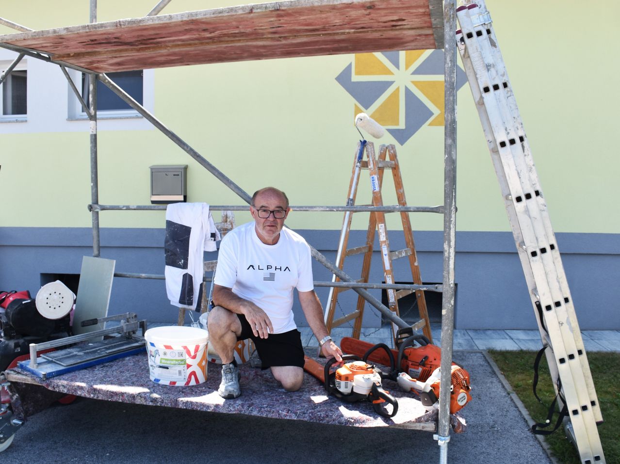 Ein Mann in einem weißen Alpha-Shirt und schwarzen Shorts kniet auf einem Gerüst, hält einen Farbroller, mit einem Eimer, Werkzeugen und einer Leiter in der Nähe.