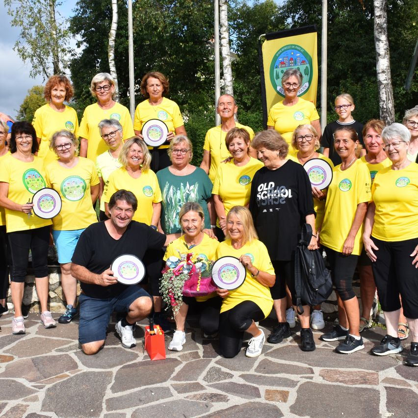 Eine Gruppe von Menschen in gelben T-Shirts posiert für ein Foto, einige halten Teller, in einer Außenumgebung mit Bäumen und einem Banner im Hintergrund.
