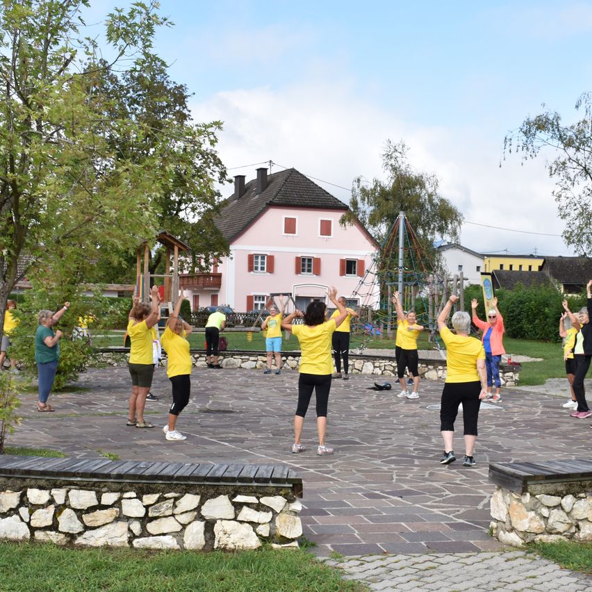 Eine Gruppe von Menschen in gelben Shirts und schwarzen Hosen macht Outdoor-Übungen in einem Park mit einem rosa Haus im Hintergrund.