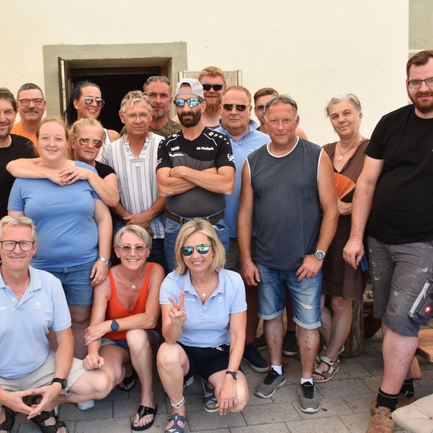 Eine Gruppe von Menschen mit Sonnenbrillen und Hüten posiert für ein Foto vor einem Gebäude. Sie alle lächeln.