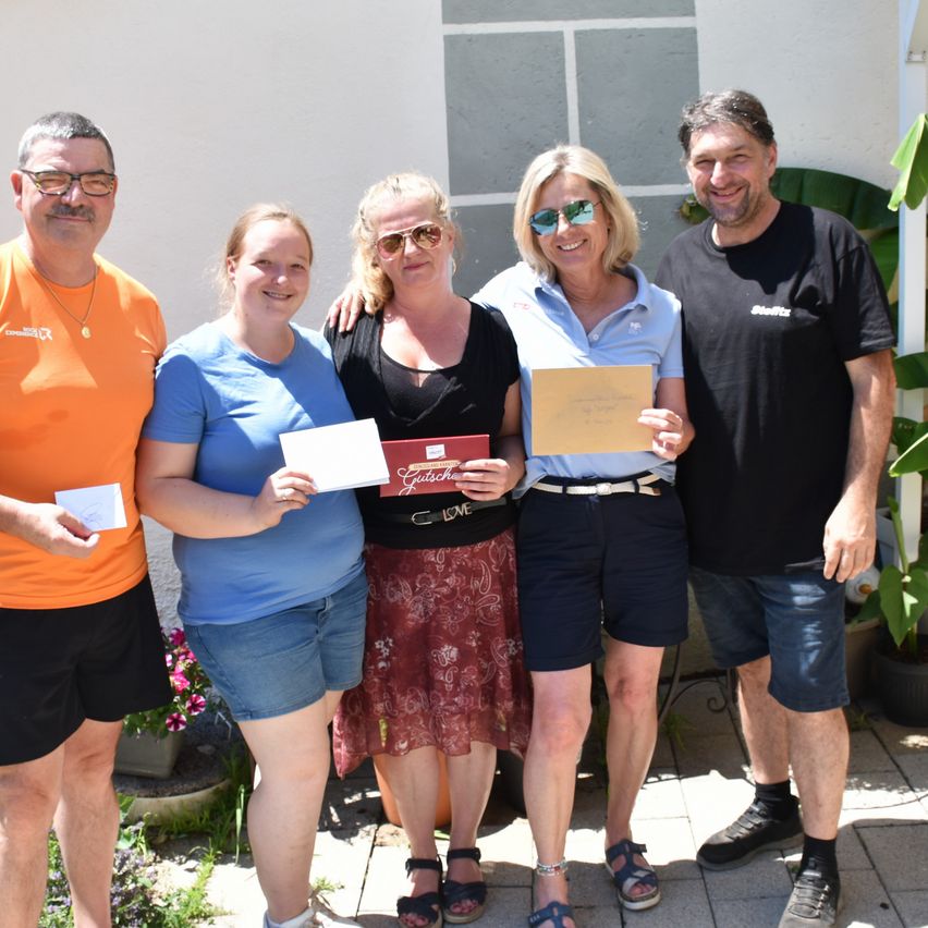 Fünf Personen posieren für ein Foto im Garten mit Urkunden. Eine Frau hält eine rote Urkunde, und zwei Frauen halten weiße Papiere. Ein Mann lächelt rechts.