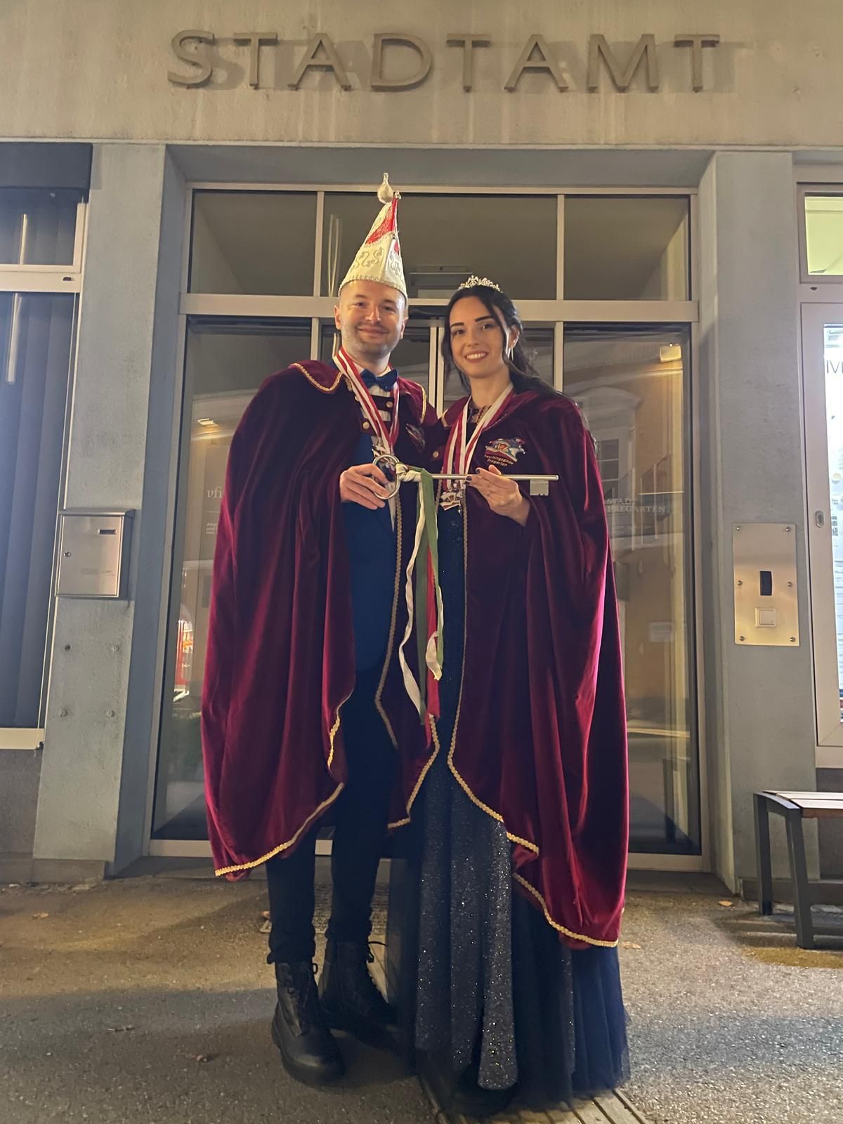 A man and a woman in costumes stand in front of a building. The man wears a red cape, hat, and bow tie. The woman wears a red cape and a crown. They hold a stick.