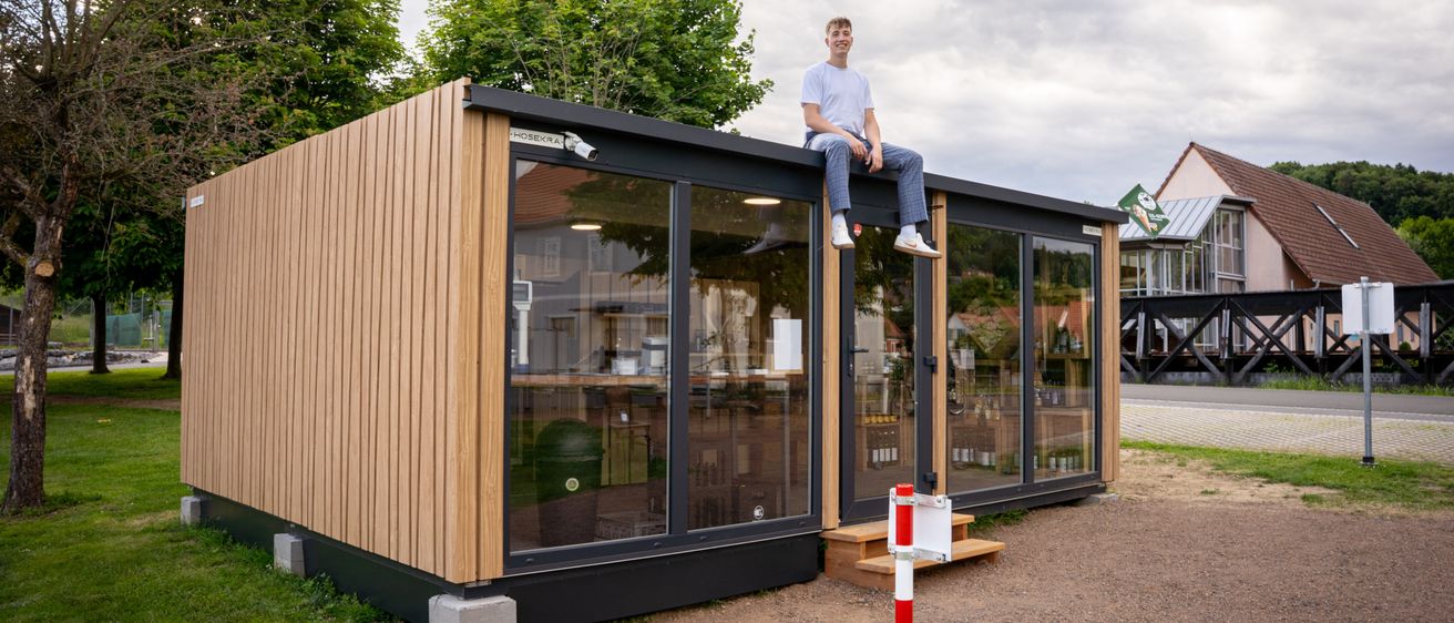 Ein Mann sitzt auf dem Dach eines modernen, umweltfreundlichen Gebäudes mit großen Fenstern, umgeben von Grün und einem bewölkten Himmel.