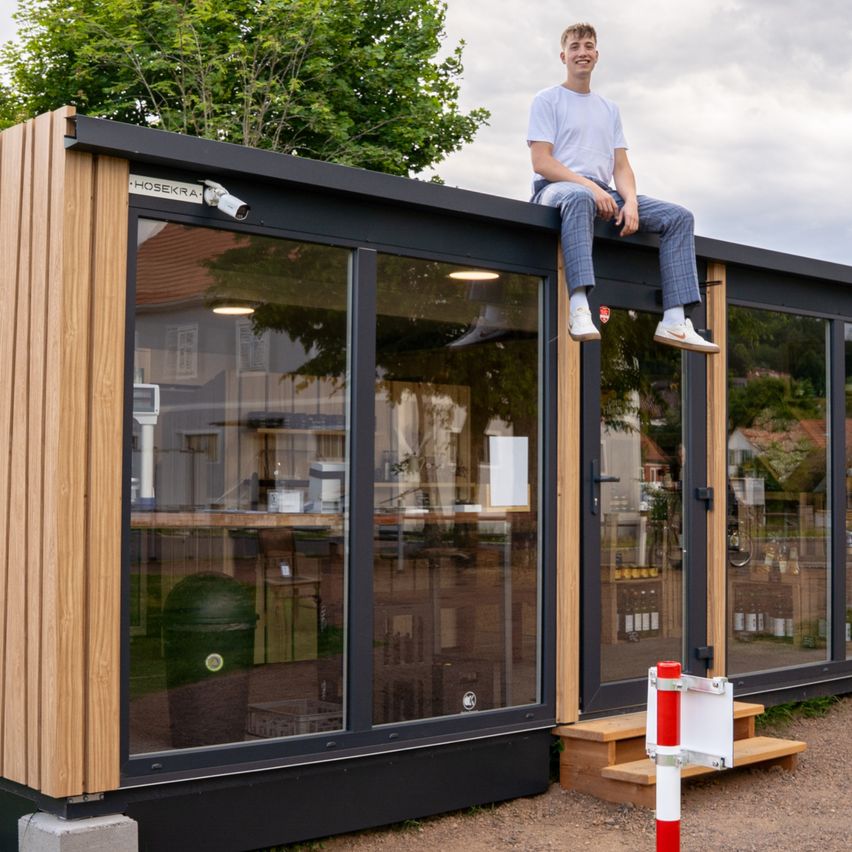 Ein Mann sitzt auf dem Dach eines modernen, umweltfreundlichen Gebäudes mit großen Fenstern, umgeben von Grün und einem bewölkten Himmel.