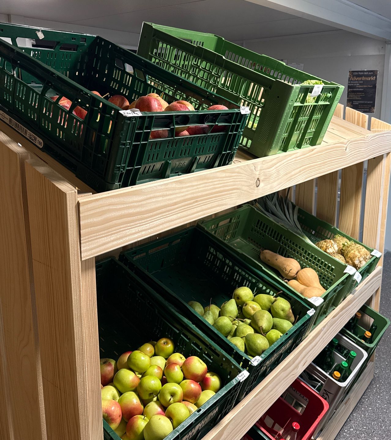 Ein hölzerne Regal mit grünen Körben enthält verschiedene Obst und Gemüse. Äpfel, Birnen und Kürbis sind sichtbar. Das oberste Regal hat zwei Körbe mit Äpfeln. Die unteren Regale haben grüne Körbe mit Birnen und Kürbis.