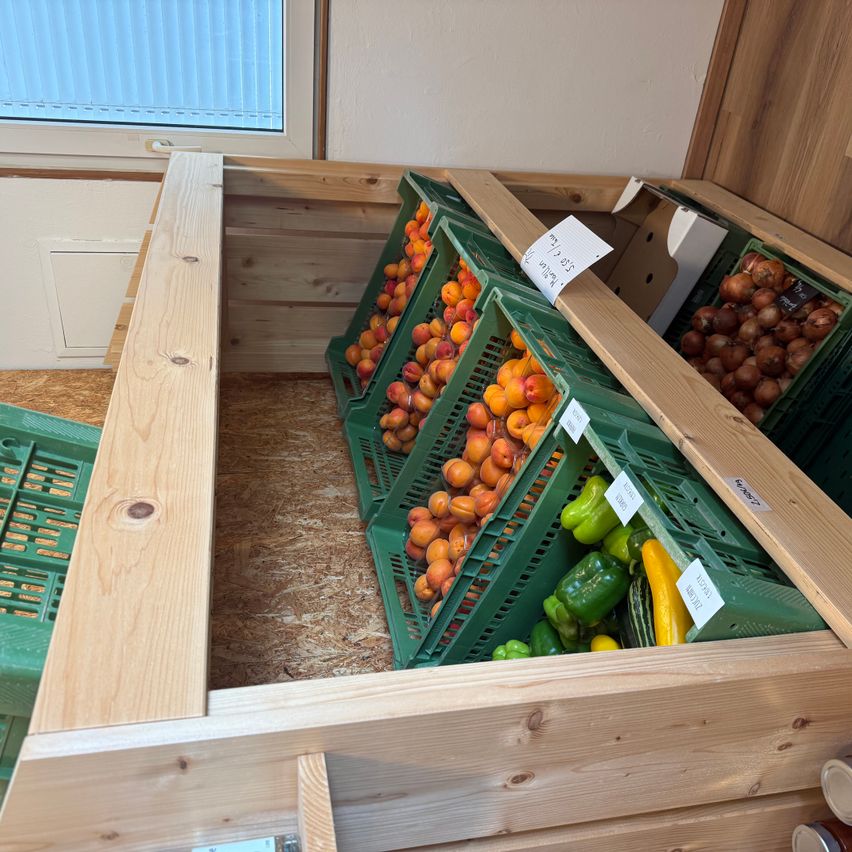 Ein Holzregal zeigt grüne Kisten gefüllt mit Pfirsichen, Paprika und Zwiebeln, positioniert in einem Raum mit einem Fenster.