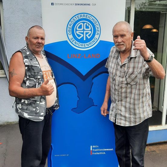 Zwei Männer stehen vor einem blauen Banner, einer zeigt den Daumen hoch und der andere lächelt. Sie tragen beide Freizeitkleidung.