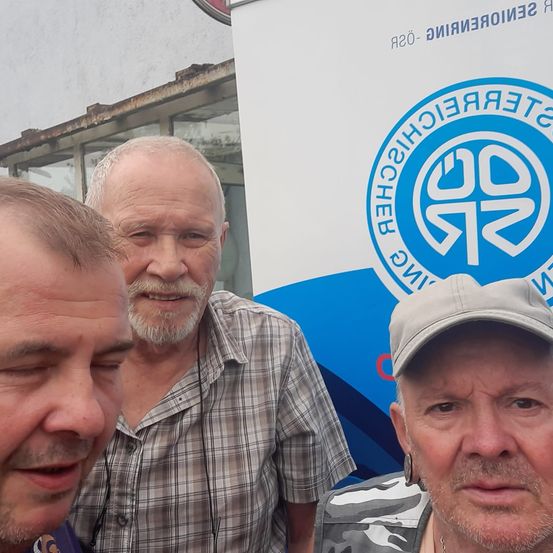 Drei Männer stehen vor einem Gebäude. Der Mann in der Mitte trägt ein kariertes Hemd, während der Mann auf der linken Seite eine graue Mütze trägt.