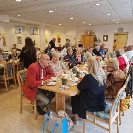 Eine Gruppe von Menschen, einschließlich Männer und Frauen, sitzen um einen Esstisch in einem Raum herum, möglicherweise bei einer Zusammenkunft. Der Tisch ist mit Tellern, Tassen und Essen gedeckt.