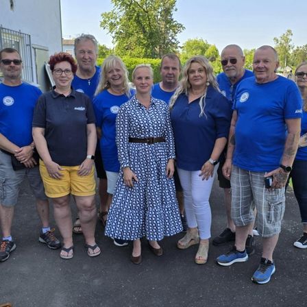 Eine Gruppe von Erwachsenen, die Blau tragen, posiert für ein Foto. Einige von ihnen tragen Brillen. Die Frau in der Mitte trägt ein Kleid. Der Hintergrund ist unscharf, mit einem Gebäude, Bäumen und einem Auto.