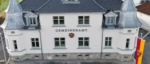 Luftaufnahme eines weißen Gebäudes mit grauem Dach, beschriftet mit Gemeindeamt, mit einem Fahnenmast und einer Flagge, umgeben von einem Rasen und einem Ziegelweg.