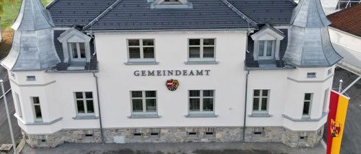Luftaufnahme eines weißen Gebäudes mit grauem Dach, beschriftet mit Gemeindeamt, mit einem Fahnenmast und einer Flagge, umgeben von einem Rasen und einem Ziegelweg.