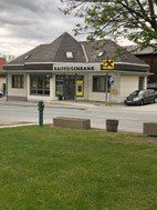 Ein weißes Gebäude mit grauem Dach, mit der Aufschrift Raiffeisenbank, befindet sich an der Seite einer Straße. Es gibt Bänke, ein Auto und einen Rasenbereich. Ein Baum steht am Straßenrand.