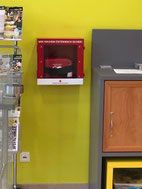 Ein Raum mit gelber Wand verfügt über einen montierten roten Feuerlöscherkasten, ein Metallregal mit Gegenständen, eine Steckdose und einen grauen Schrank mit einer hölzernen Schublade.
